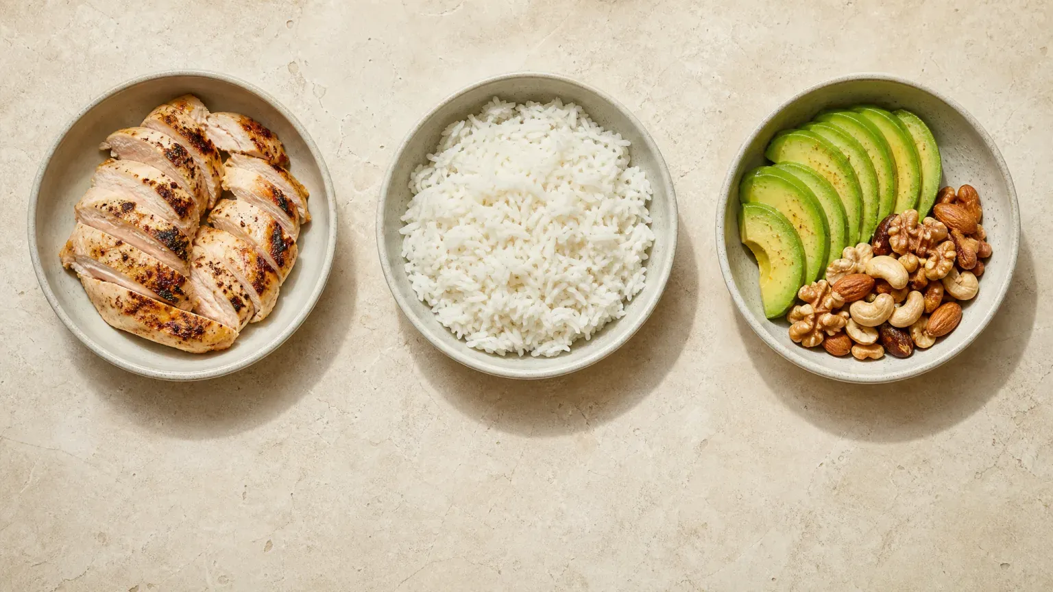 Three pale ceramic bowls in a row on stone, holding sliced grilled chicken, cooked white rice, and avocado with mixed nuts